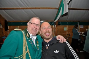 Fröhliche Männer bei Schützenfest, einer trägt Uniform mit Abzeichen, andere macht Spaß, in Zelt, festliche Stimmung, Gemeinschaft, Freundschaft, Karnevalsfeier.