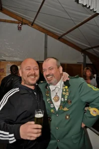 Fröhliche Schützenbrüder in traditioneller Uniform mit zahlreichen Abzeichen bei einem Fest im Zelt, elegant und gesellig, mit Bier in der Hand, in Köln.