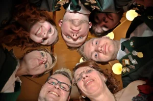 Fröhliche Schützenbrüder und -schwestern in traditioneller Kleidung beim Gruppenfoto, Blick nach unten, lachend, in festlicher Umgebung, symbolisiert Gemeinschaft und Vereinsgeist.