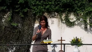 Frau steht mit Mikrofon und Buch in der Hand, während sie spricht. Im Hintergrund ist ein Kreuz sichtbar.