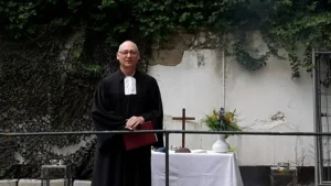 Ein Pfarrer in schwarzer Robe steht vor einem Tisch mit Blumen und einem Kreuz im Hintergrund.